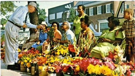 Old postcard image depicting a man, Robert B. House, former chancellor of UNC-Chapel Hill, buying flowers from a group of African-American florists known as the Flower Ladies on Franklin Street in Chapel Hill.