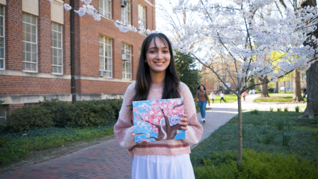 A student, Daneen Khan, posing with a painting she made while standing on a brick pathway next to a building on the campus of UNC-Chapel Hill.